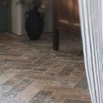 Podłoga z trawertynu ASH MIX 30,5x10x1 cm w ułożeniu jodełkowym, harmonijnie komponująca się z drewnianymi meblami i zasłoną w paski.